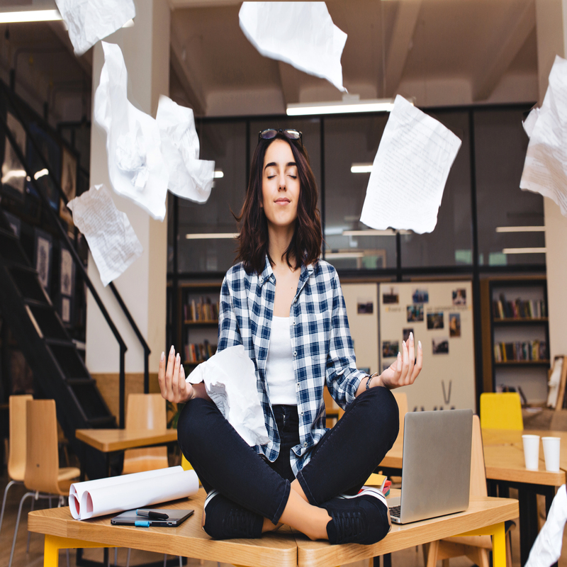 Young pretty joyful brunette woman meditating on table surround work stuff and flying papers. Cheerful mood, taking a break, working, studying, relaxation, true emotions.